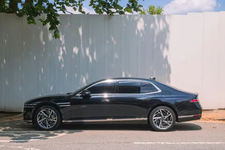 Black Genesis G90 luxury sedan parked in side profile on a street in front of a white fence with tree shadows.