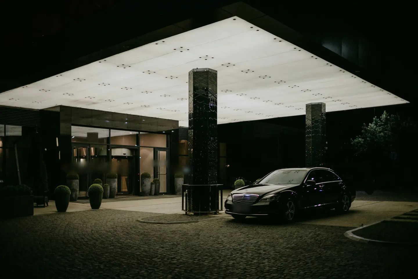 Black Mercedes-Benz S-Class sedan waiting under the lit entrance canopy of a modern building at night.