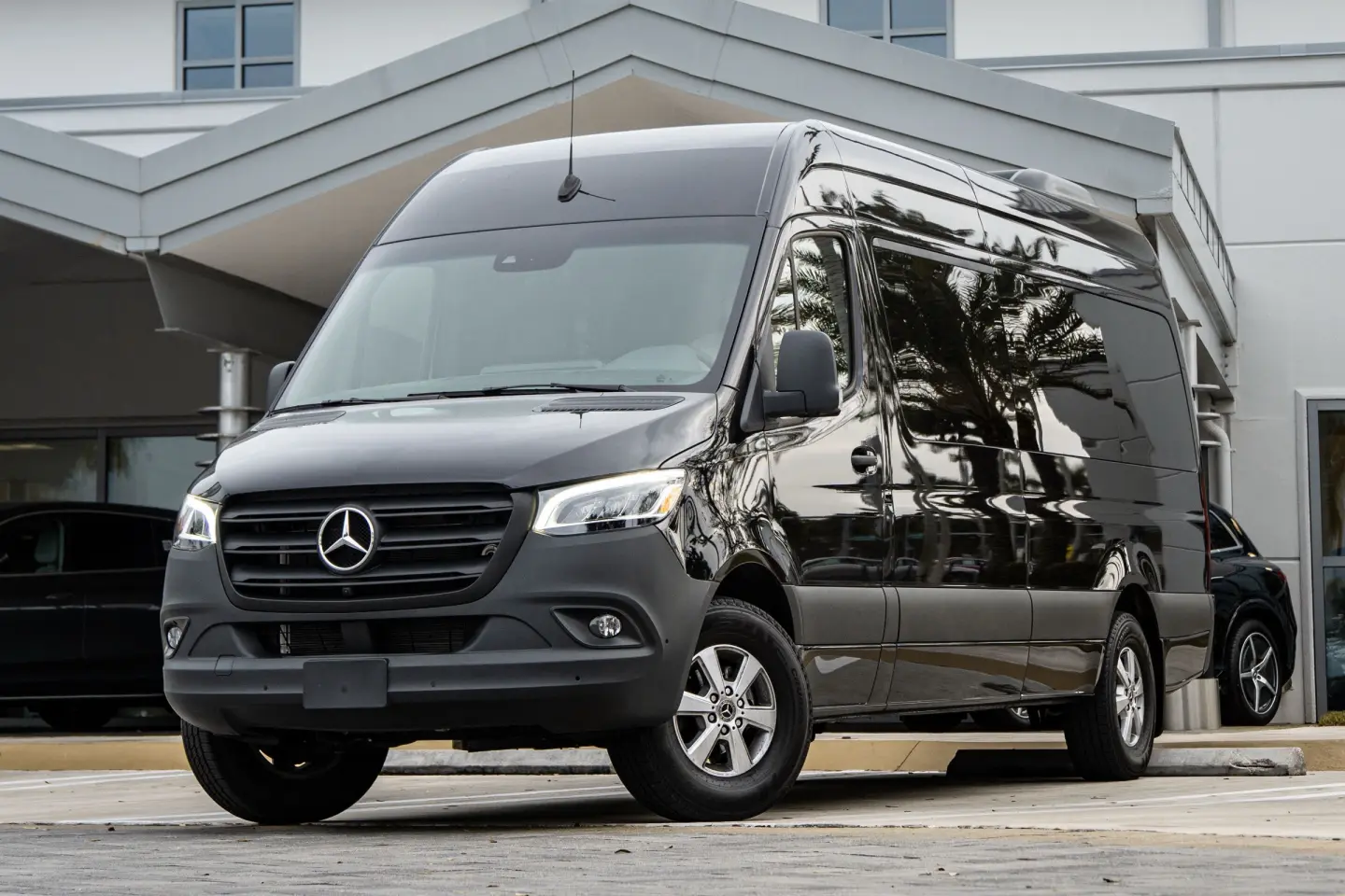 Black high-roof Mercedes-Benz Sprinter passenger van with tinted windows, parked under a building canopy.
