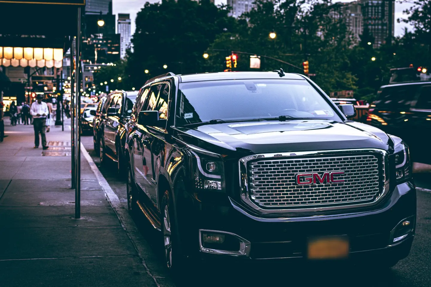 Black GMC Yukon Denali SUV waiting at a city curb at dusk, with streetlights and traffic signals glowing behind.