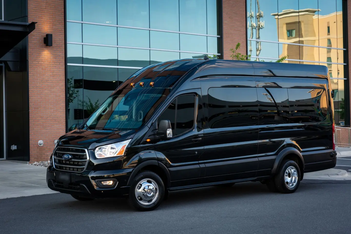 Black high-roof Ford Transit passenger van with dual rear wheels parked outside a glass and brick building.
