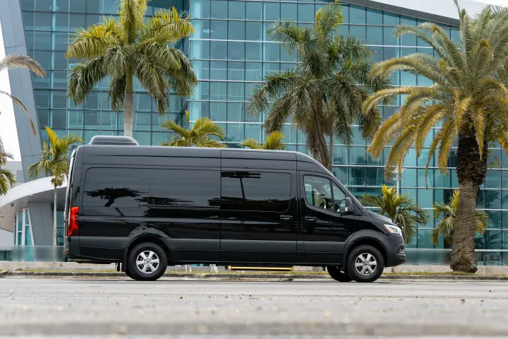 Black high-roof Mercedes-Benz Sprinter van with blacked-out windows in side profile, by a glass building and palm trees.