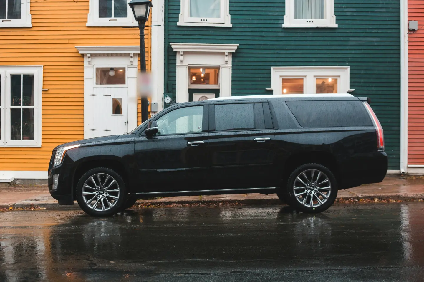 Black Cadillac Escalade ESV parked in side profile on a rain-wet street in front of colorful wooden row houses.