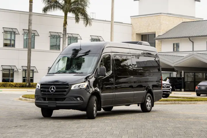 Black Mercedes-Benz Sprinter passenger van in front three-quarter view on a paver forecourt outside a white building.