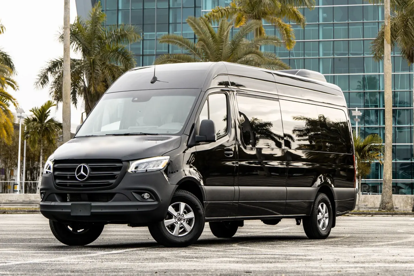 Black high-roof Mercedes-Benz Sprinter van in front three-quarter view, parked by a glass building and palm trees.