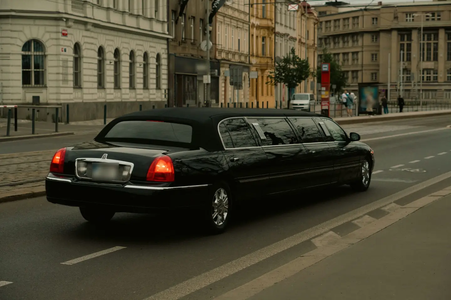 Black Lincoln Town Car stretch limousine driving along a city street past historic stone buildings, seen from behind.