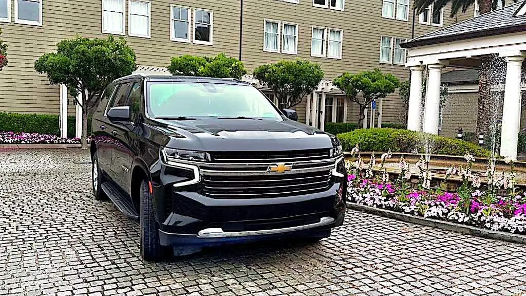 Front view of a black Chevrolet Suburban SUV at a hotel entrance, with flower beds and a fountain behind.