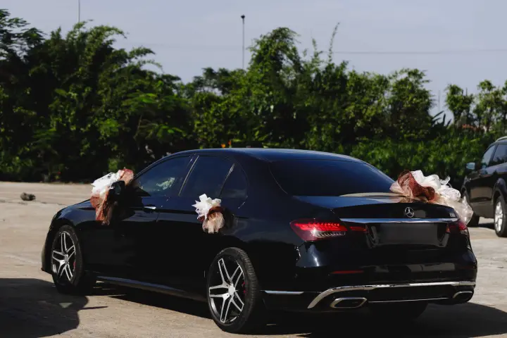 Black Mercedes-Benz E-Class wedding car decorated with cream and copper-brown tulle bows, parked in front of green trees.