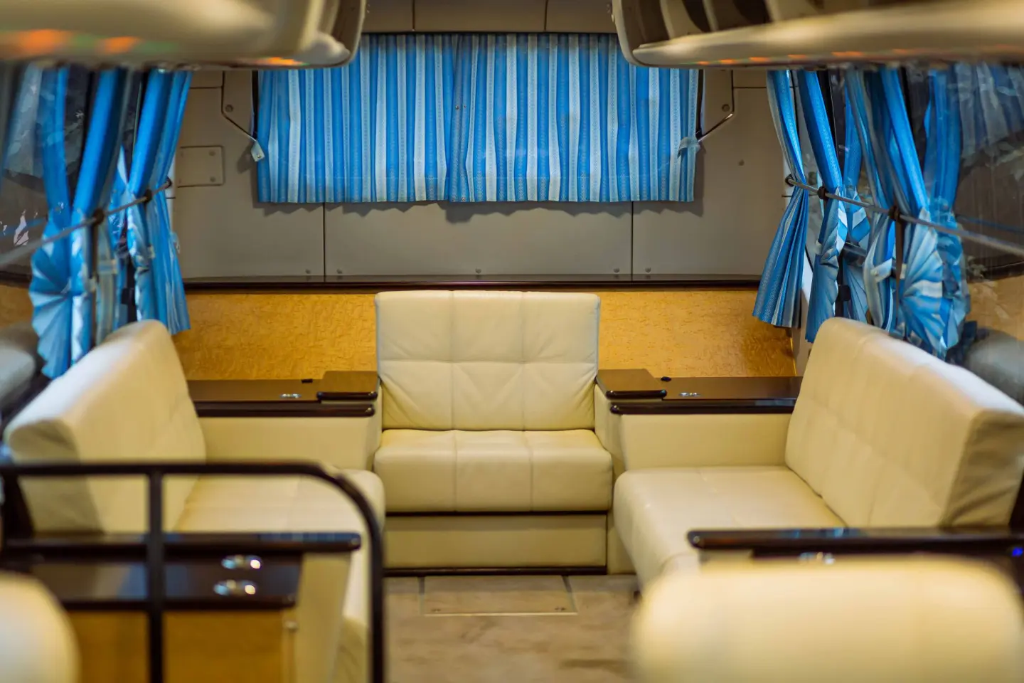 Cream leather lounge sofas and wood side tables inside a charter bus, with blue striped curtains at the windows.