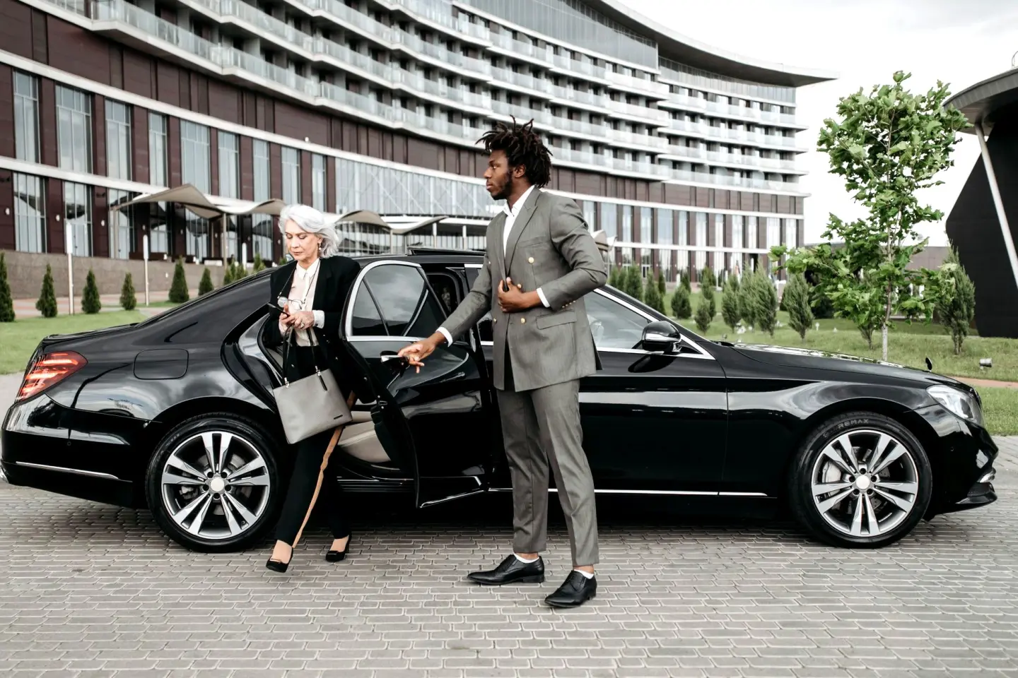 Chauffeur in a gray suit holds the rear door of a black Mercedes-Benz S-Class as a business traveler steps out.