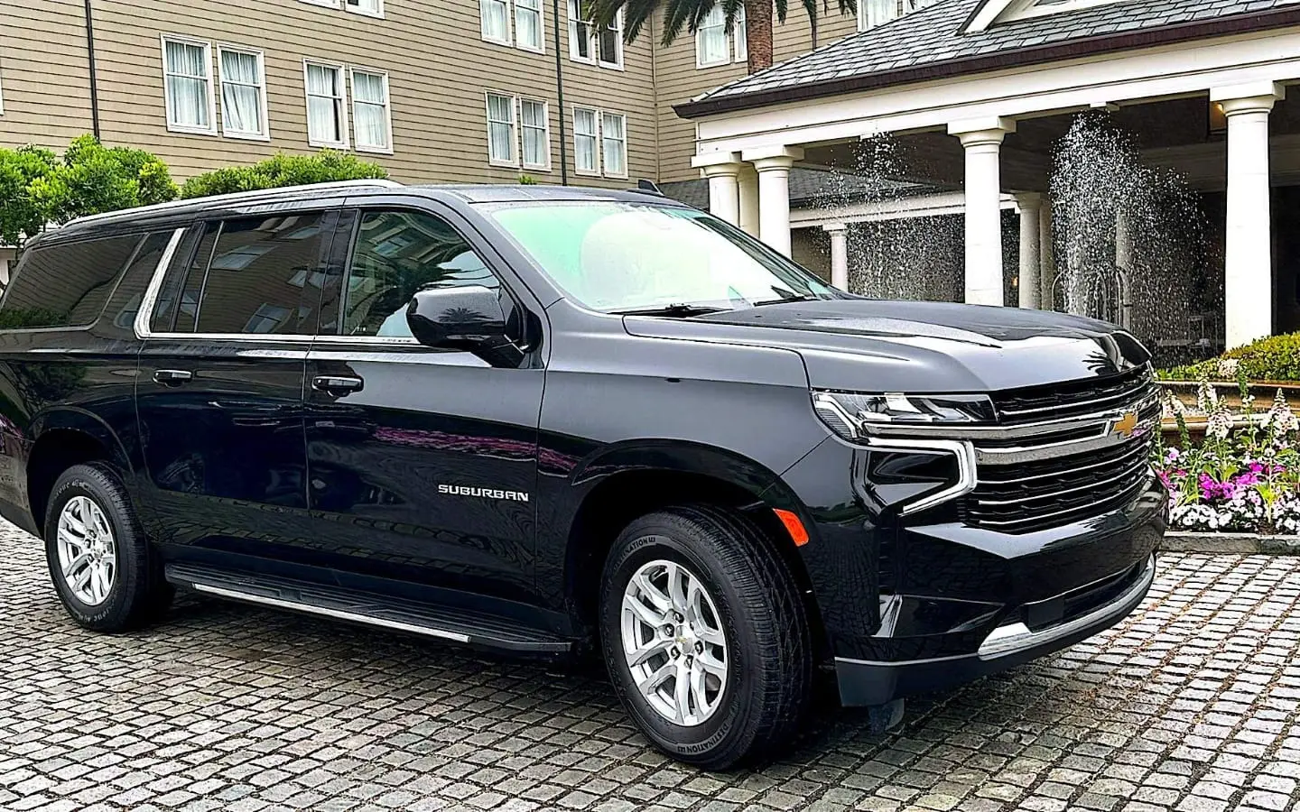 Black Chevrolet Suburban SUV parked on a cobblestone hotel forecourt beside a fountain and flower beds.