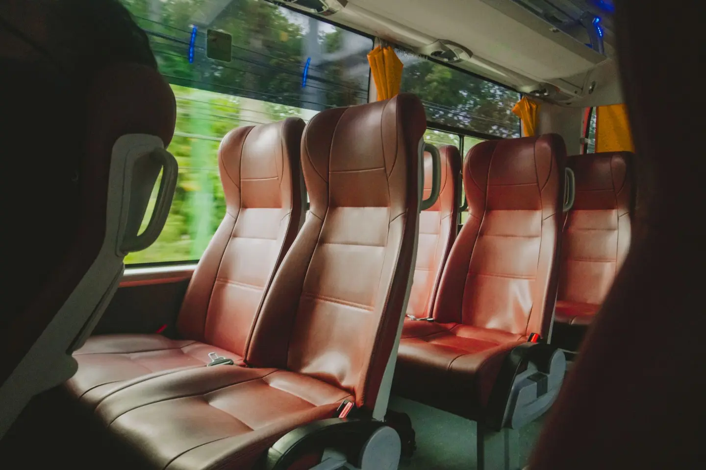Rows of brown leather high-back seats beside the windows inside a coach bus, with yellow curtains tied back.