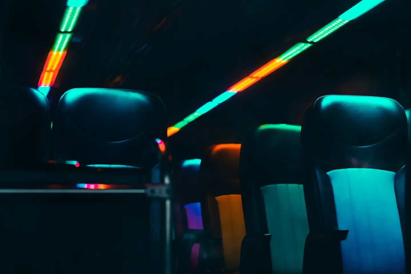 Dark vehicle cabin with high-back seats lit by green, orange and blue LED strip lights along the ceiling.