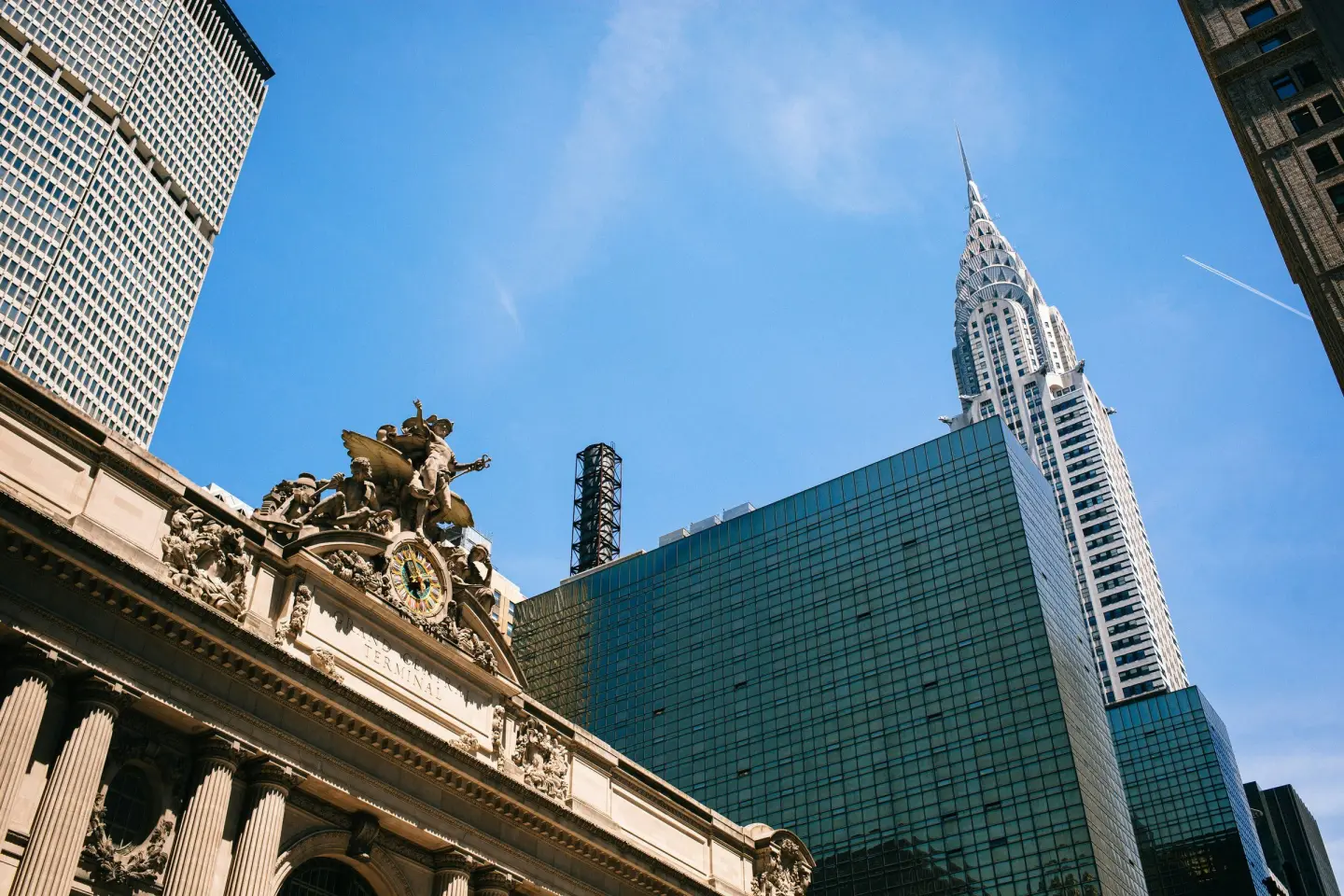 Low-angle view of the Grand Central Terminal clock and sculpture group, with the Chrysler Building spire against a blue sky.