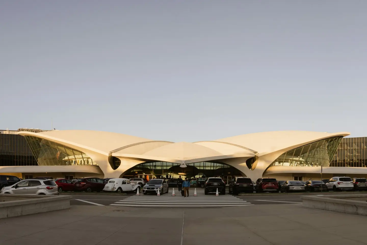 Cars and dark SUVs lined up in front of the curved concrete wings of the TWA Flight Center at JFK Airport.