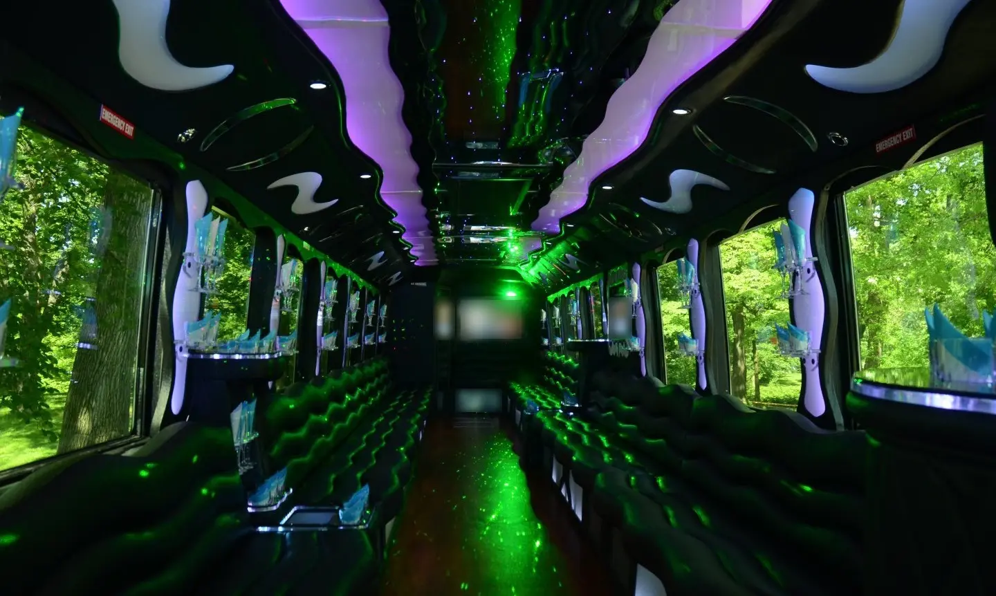 Long party bus cabin with black leather perimeter seating, green laser lights, a purple-lit ceiling and glassware racks.