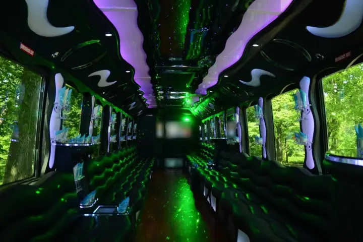 Long party bus cabin with black leather perimeter seating, green laser lights, a purple-lit ceiling and glassware racks.