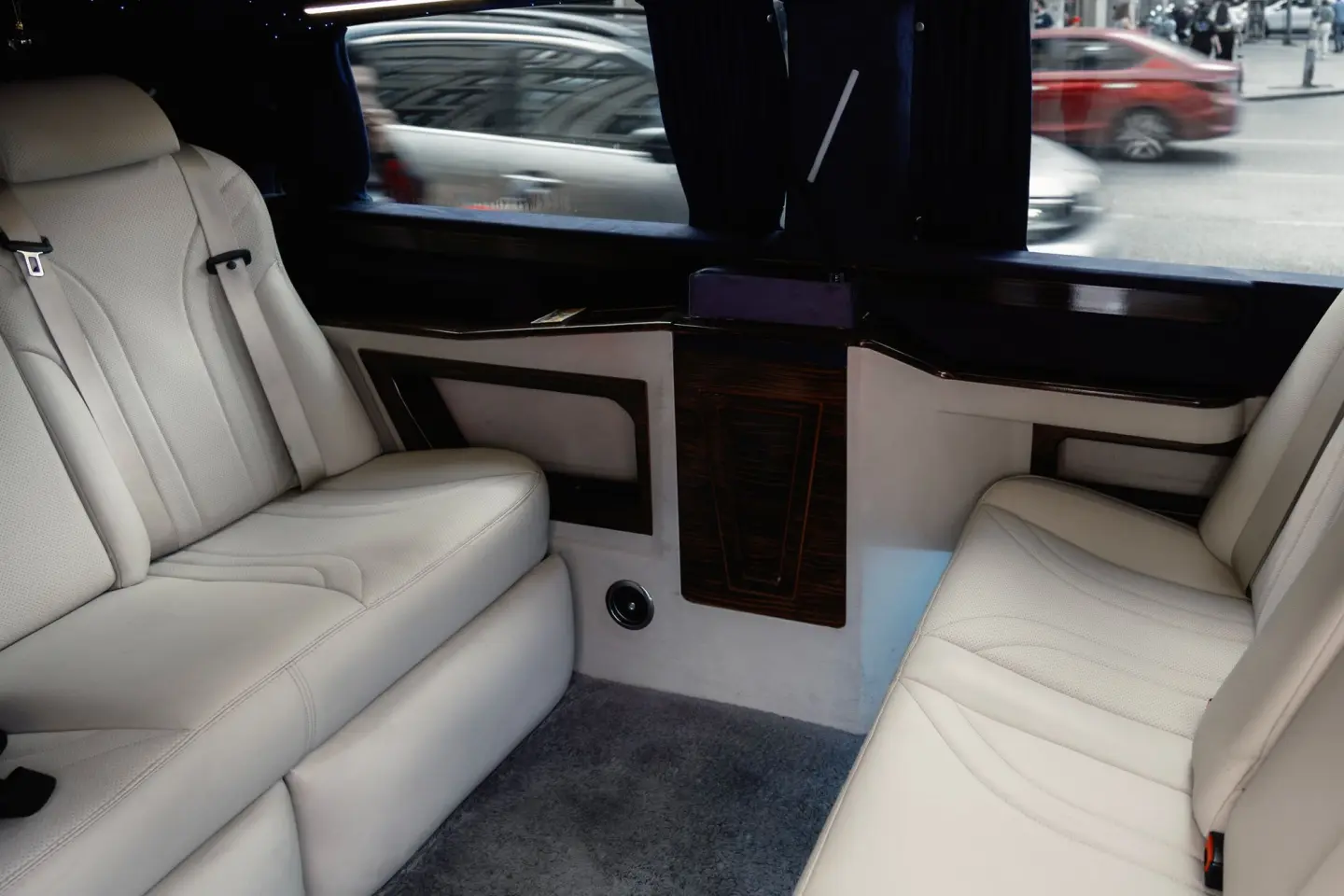 Limousine cabin with facing cream-white leather seats, wood trim and dark curtains, with city traffic passing outside.