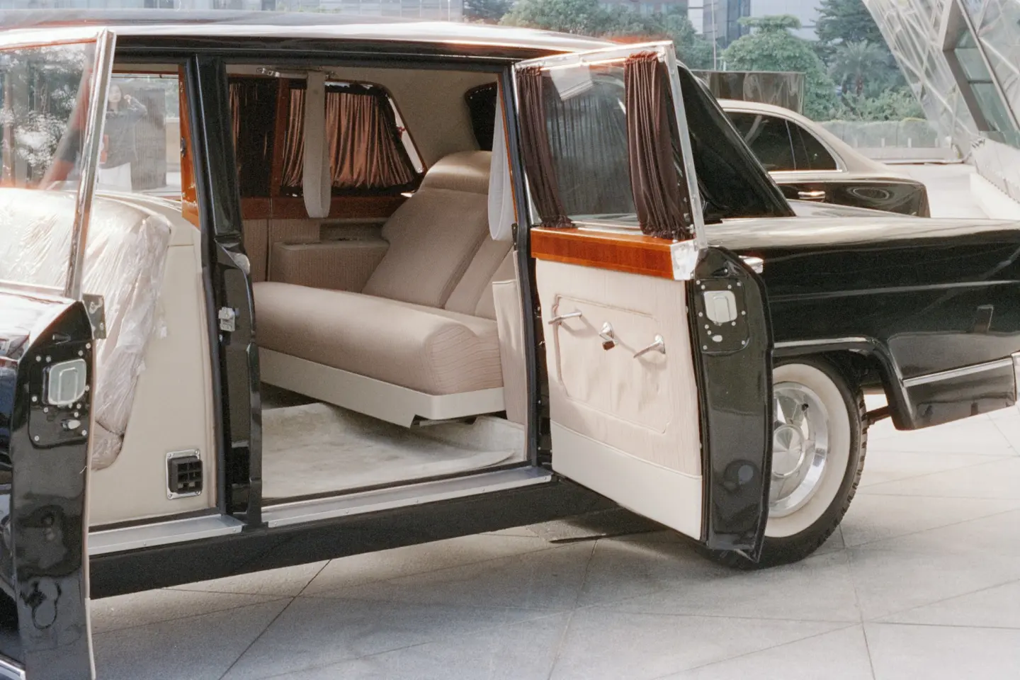 Classic black limousine in a showroom with its side doors open, showing a cream leather rear seat, curtains and wood trim.