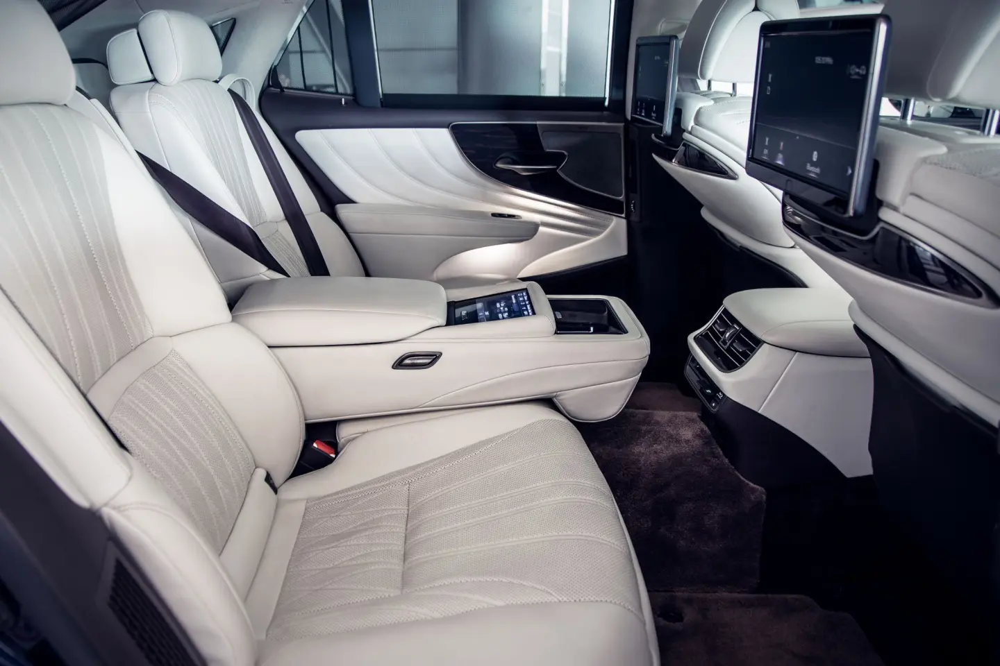 Rear seats of a luxury sedan in white leather, with a touchscreen armrest and seatback entertainment screens.