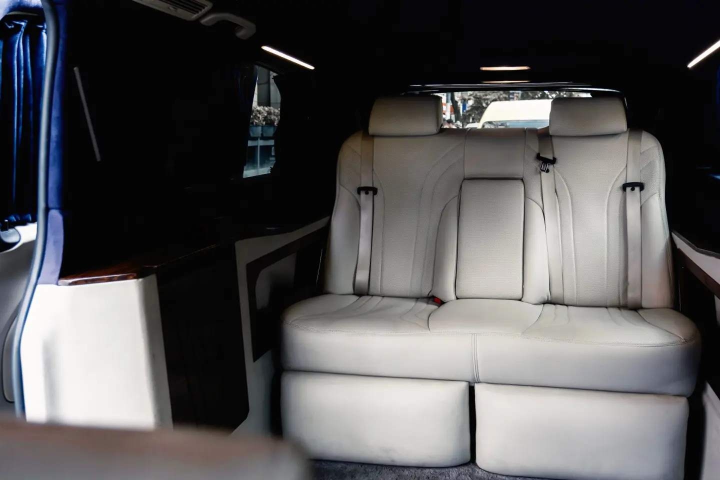 Cream leather rear bench seat in a dark luxury van cabin with wood trim, curtains and LED ceiling lights.