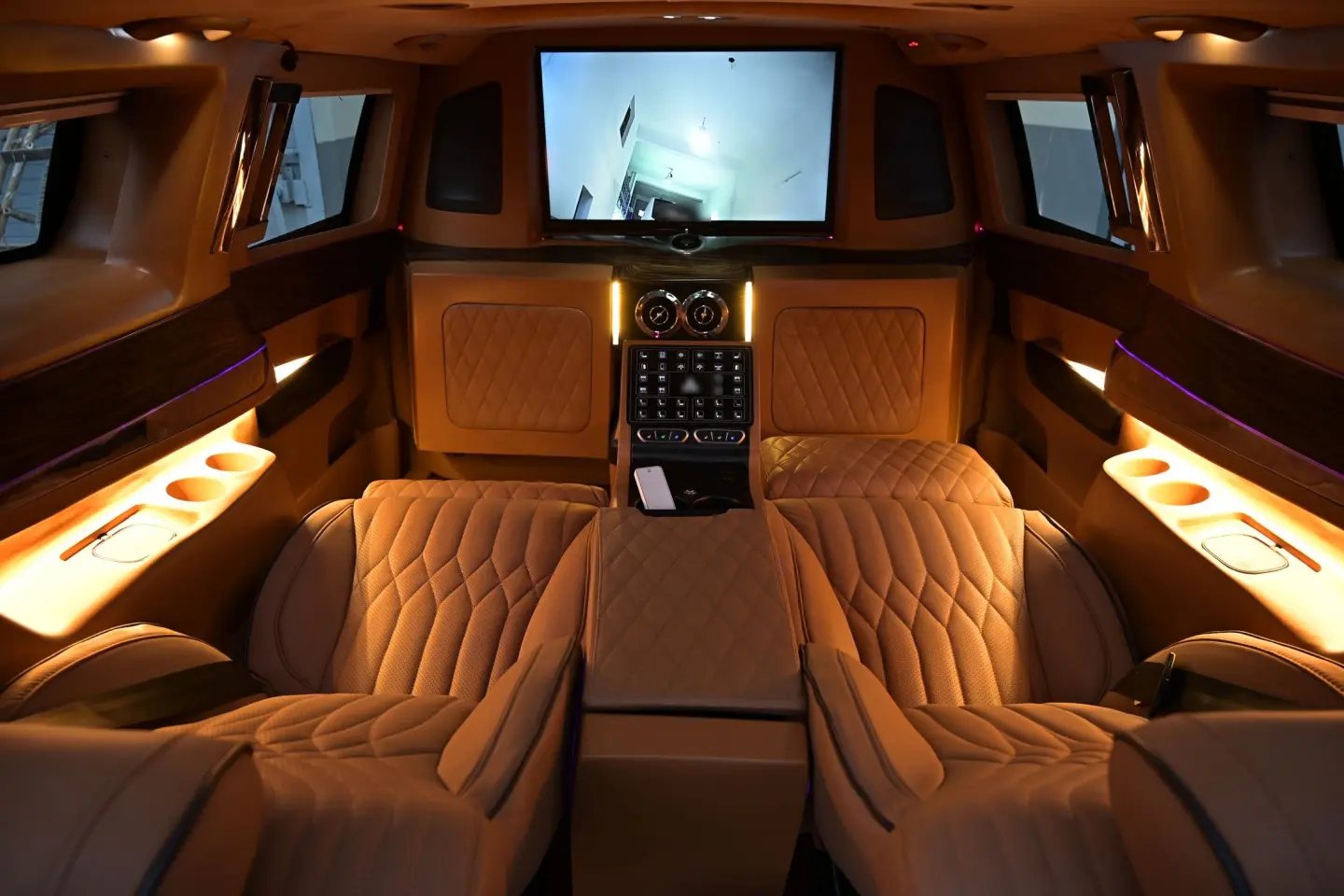 Luxury van rear cabin with quilted tan leather captain chairs, a partition-mounted TV, wood trim and warm ambient lighting.