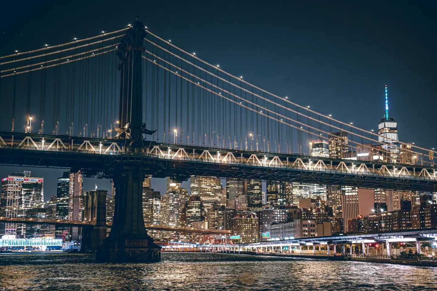 The lit Manhattan Bridge over the East River at night, with Lower Manhattan towers and One World Trade Center behind.