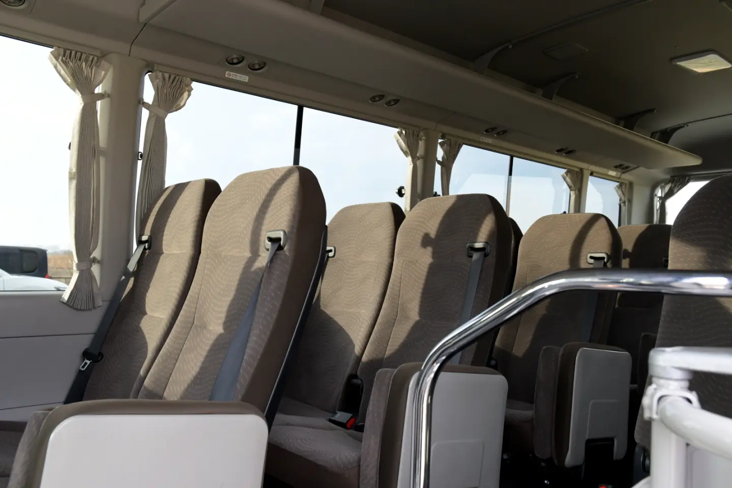 Minibus passenger cabin with taupe fabric high-back seats with seat belts, tied-back curtains and a chrome grab rail.