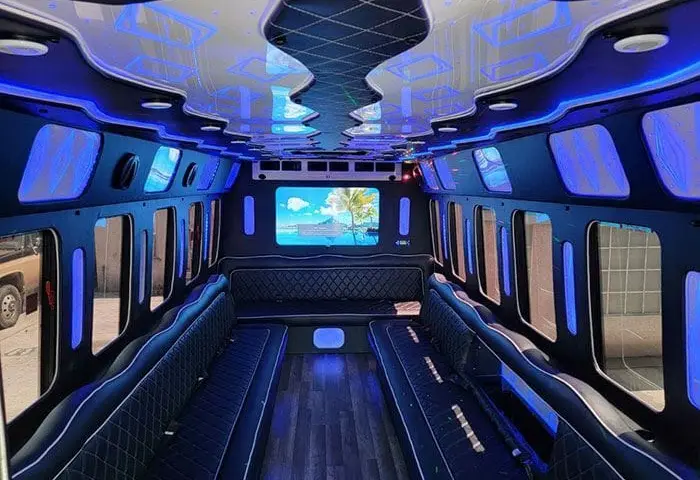 Party bus lounge with black quilted leather benches on both sides, blue LED lighting, a rear TV and a wood-look floor.