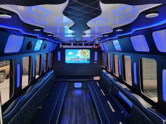 Party bus lounge with black quilted leather benches on both sides, blue LED lighting, a rear TV and a wood-look floor.