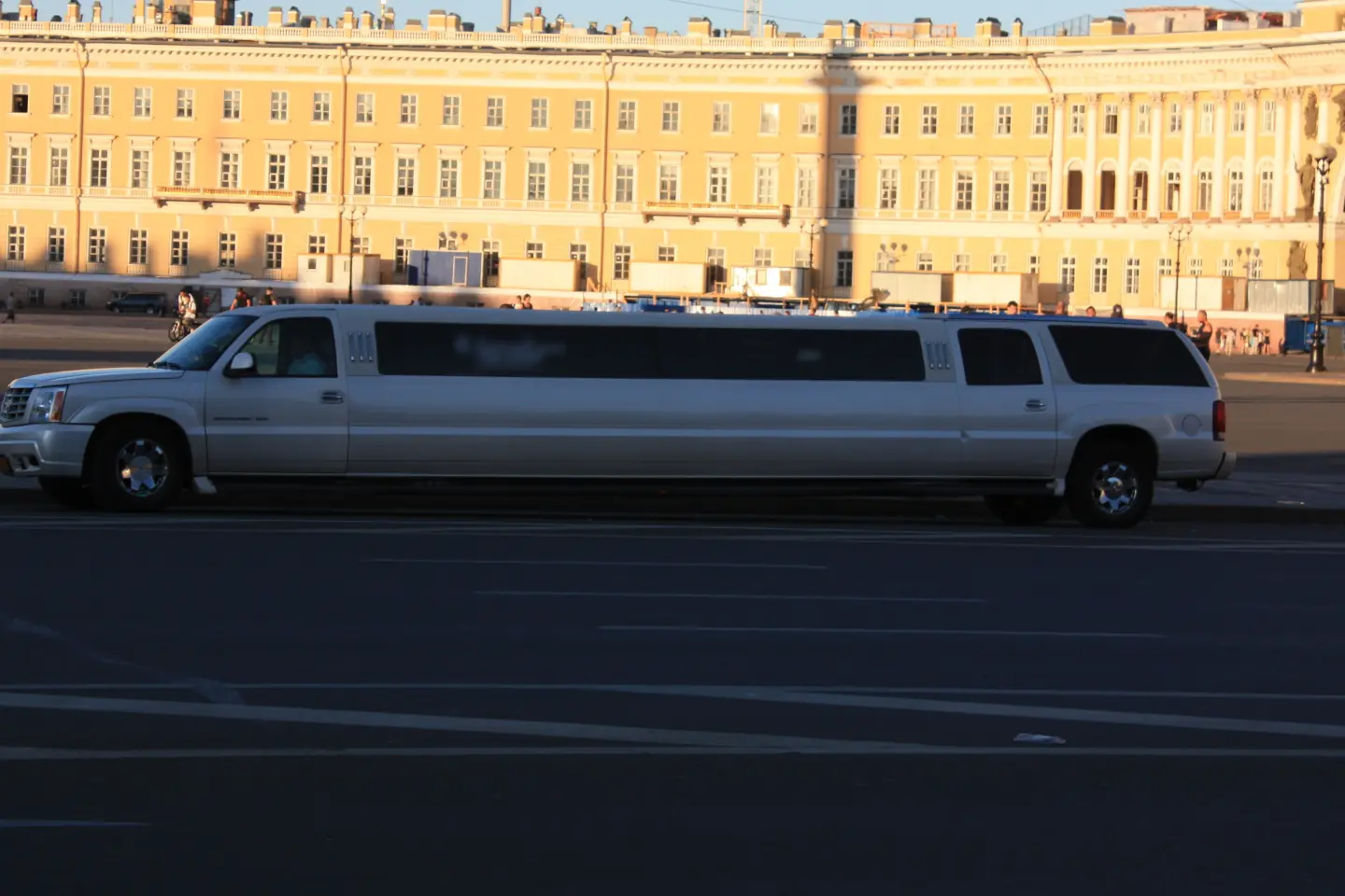 Silver Cadillac Escalade stretch limousine parked on a wide square in front of a long yellow classical building.