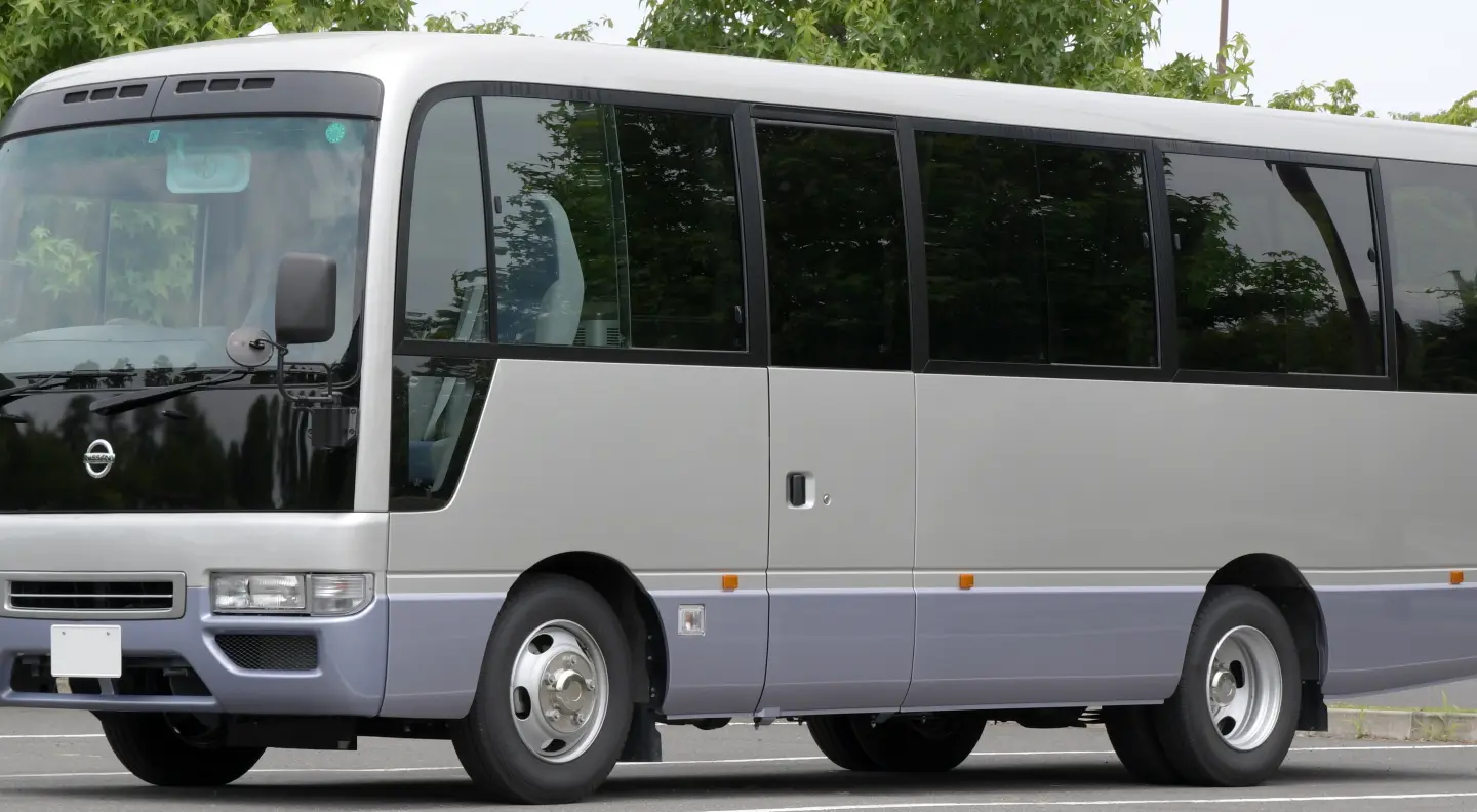 Silver and lilac mid-size minibus with a long row of tinted windows and a side passenger door, parked with trees behind.