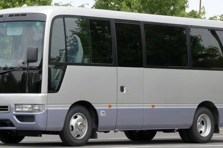 Silver and lilac mid-size minibus with a long row of tinted windows and a side passenger door, parked with trees behind.