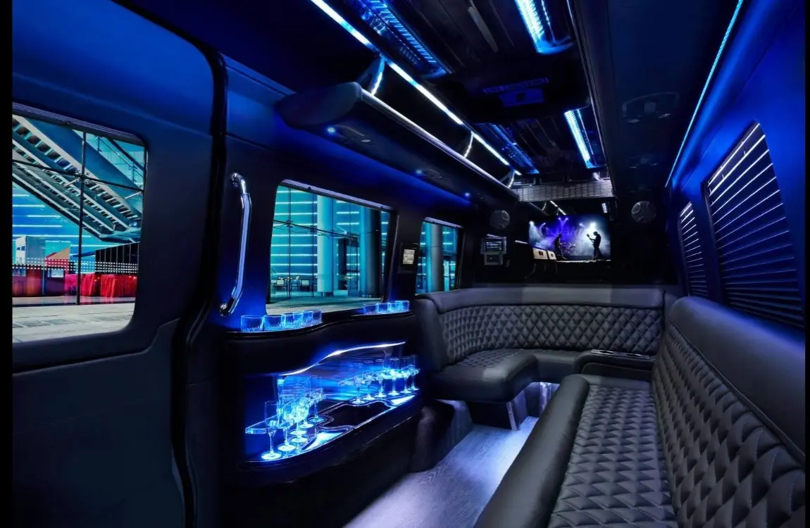 Sprinter limo interior with a glowing glassware bar, quilted black leather benches and a TV at the rear.