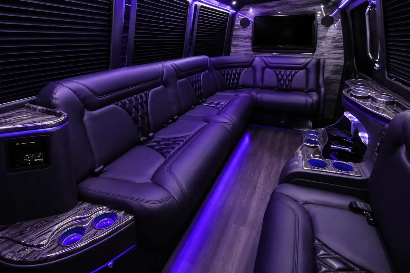 Sprinter party van lounge with purple-lit quilted leather wraparound seating, a TV and a bar console with cup holders.