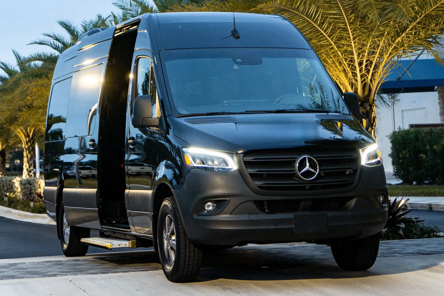 Black Mercedes-Benz Sprinter passenger van with headlights on and its sliding door open, in front of palm trees at dusk.