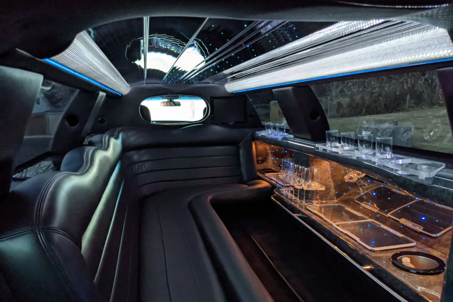 Stretch limousine interior with black leather wraparound seating, a lit bar with glassware and a mirrored starlit ceiling.