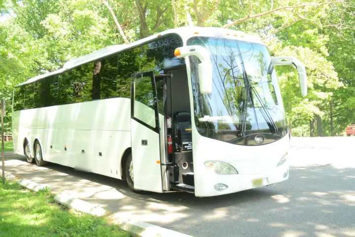 White full-size charter bus with its passenger door open, parked in a shady park lot lined with trees.