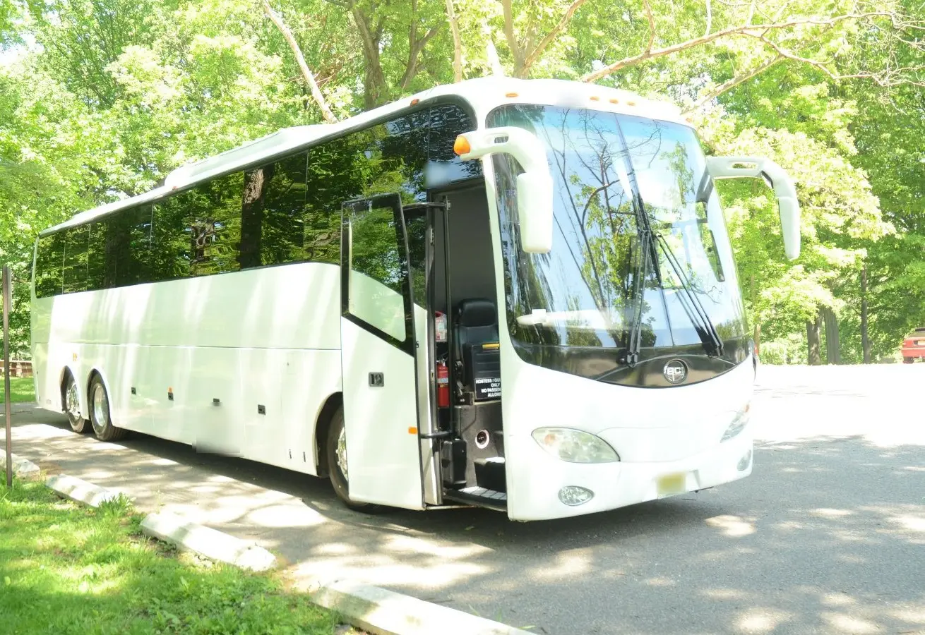 White full-size charter bus with its passenger door open, parked in a shady park lot lined with trees.