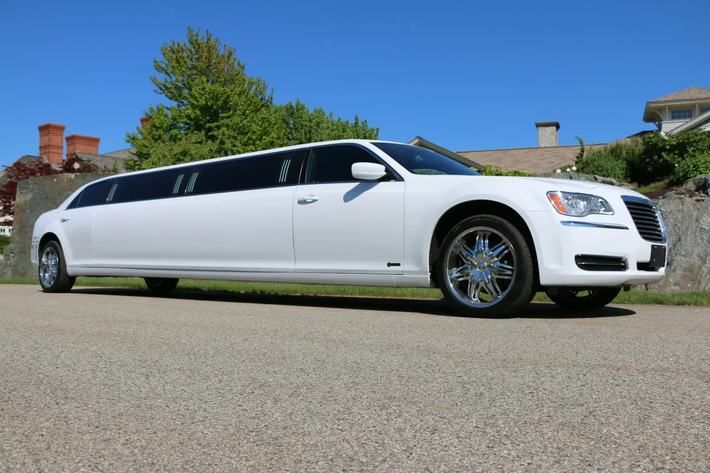 White Chrysler 300 stretch limousine with chrome wheels parked beside a lawn and stone wall under a blue sky.