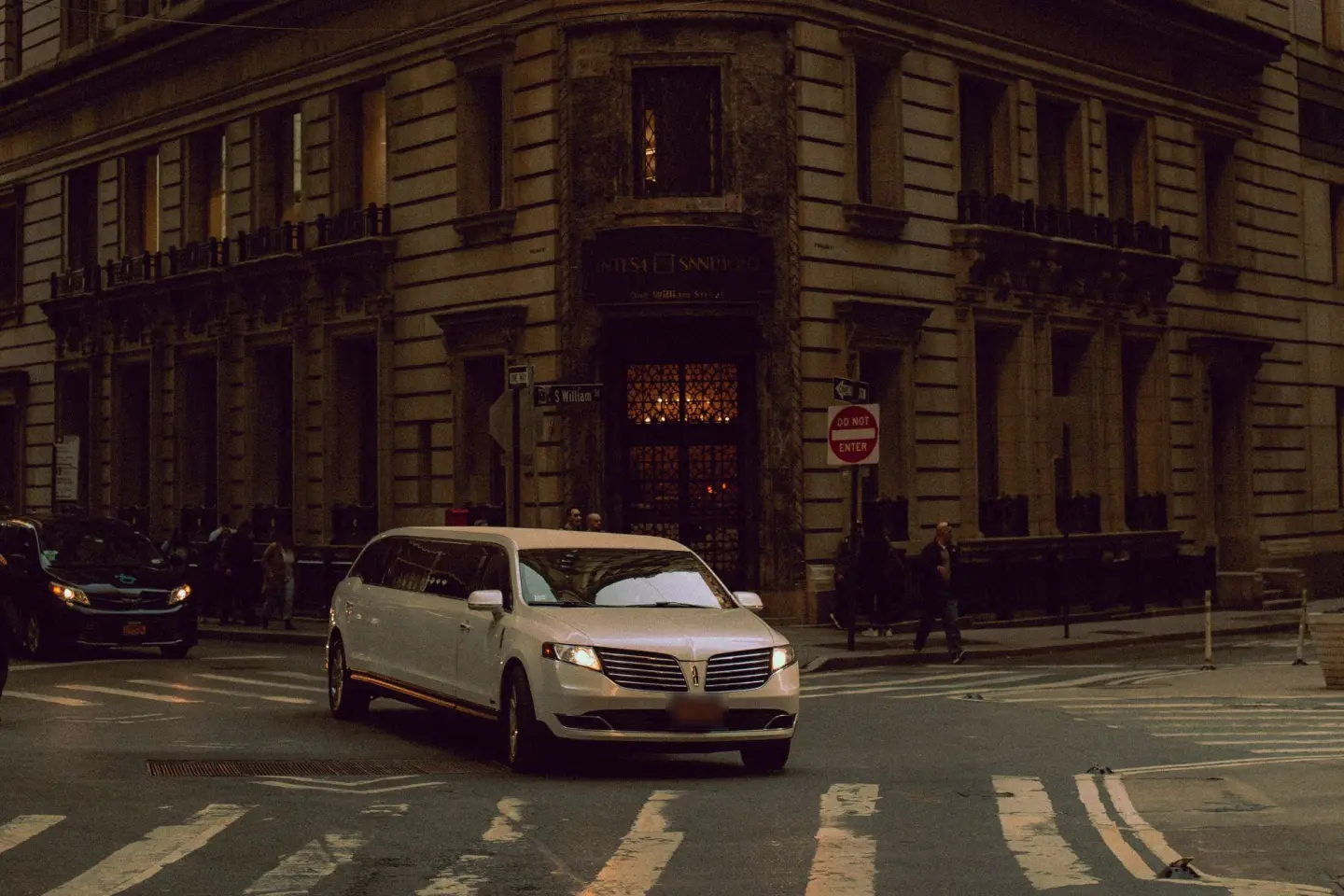 White Lincoln MKT stretch limousine turning at a city street corner beside a stone building and a do-not-enter sign.