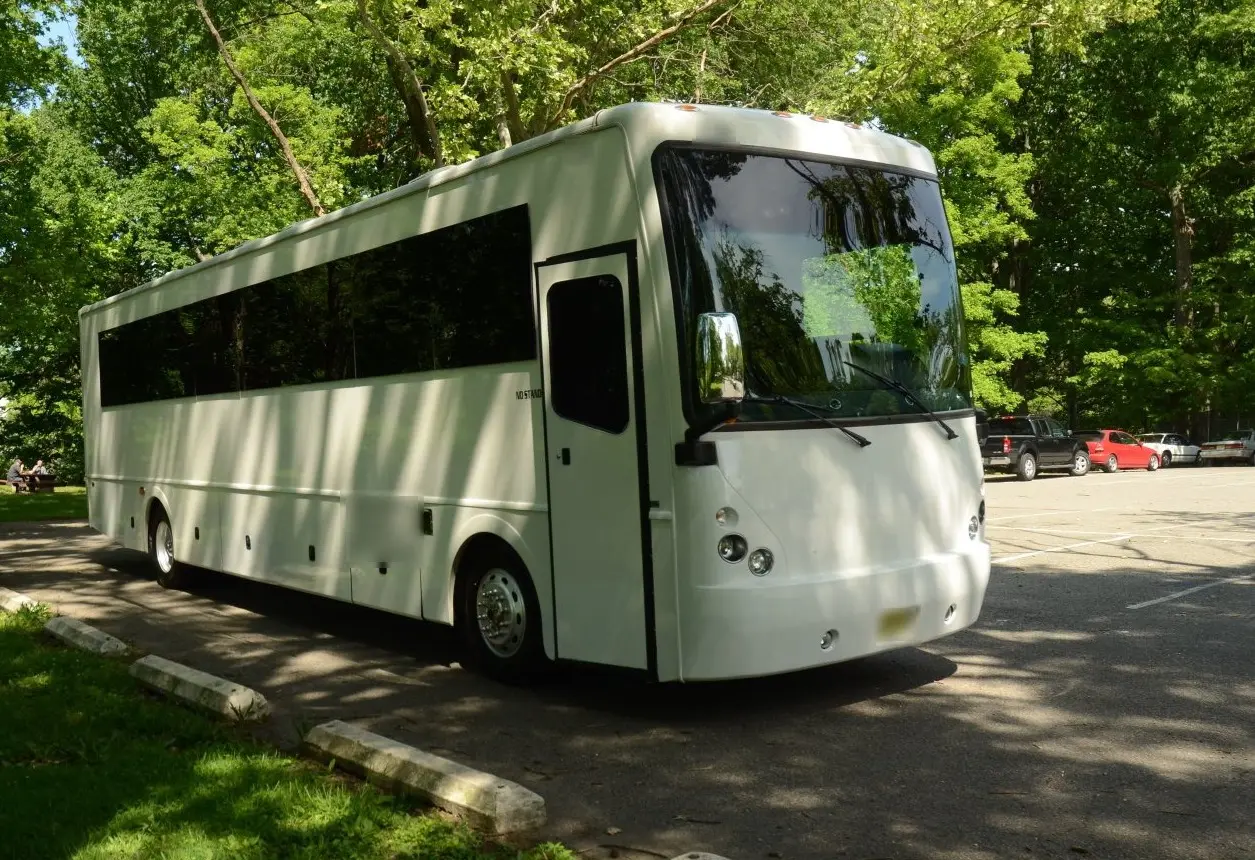 White full-size bus with a long band of tinted windows, parked under shady trees beside a park lot.